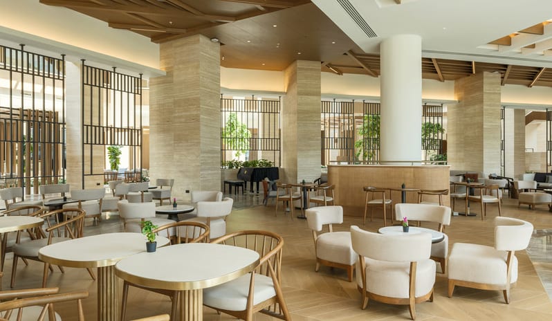 Olivae Patisserie lounge with floor-to-ceiling windows, light wood herringbone floors, and various contemporary seating arrangements under a decorative beamed ceiling at Anda Barut Collection by Kenwood Travel