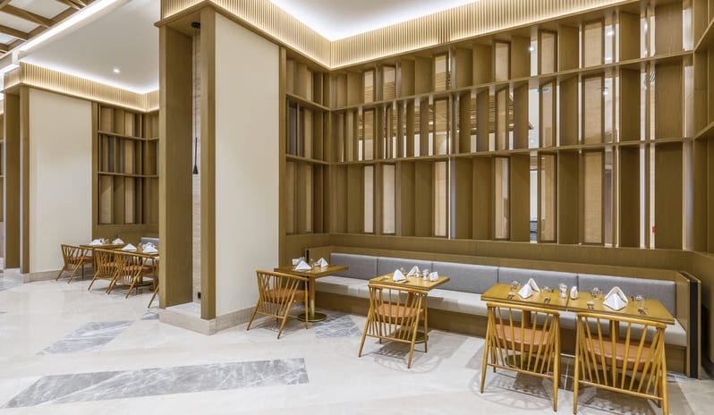 Zestful Bazaar Main Restaurant featuring long gray banquette seating, wooden dining tables, and a floor-to-ceiling decorative wood lattice wall with warm accent lighting at Anda Barut Collection by Kenwood Travel