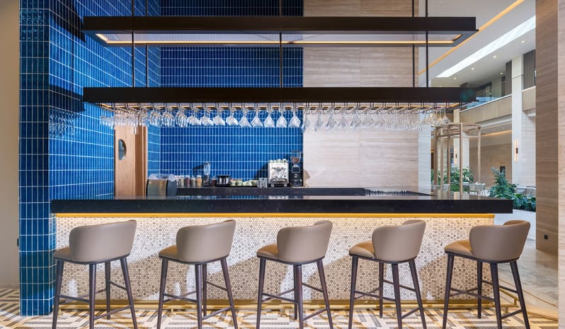 Cibes Restaurant International bar featuring a vibrant blue tiled backsplash, a black marble counter with five tan leather barstools, and an overhead rack with hanging glassware under warm LED lighting at Anda Barut Collection by Kenwood Travel