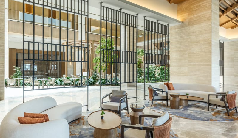 Olivae Patisserie lounge with floor-to-ceiling windows, light wood herringbone floors, and various contemporary seating arrangements under a decorative beamed ceiling at Anda Barut Collection by Kenwood Travel