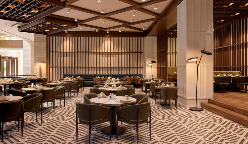 Cibes Restaurant International dining room featuring circular marble tables, olive green chairs, a distinctive geometric patterned tile floor, and a warm wood coffered ceiling at Anda Barut Collection by Kenwood Travel