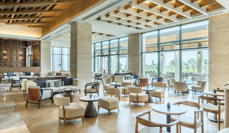 Olivae Patisserie lounge with floor-to-ceiling windows, light wood herringbone floors, and various contemporary seating arrangements under a decorative beamed ceiling at Anda Barut Collection by Kenwood Travel