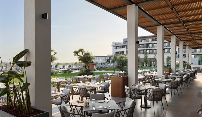 Zestful Bazaar Main restaurant terrace under a modern wooden pergola, featuring contemporary woven chairs and tables overlooking a resort garden and hotel building at Anda Barut Collection by Kenwood Travel