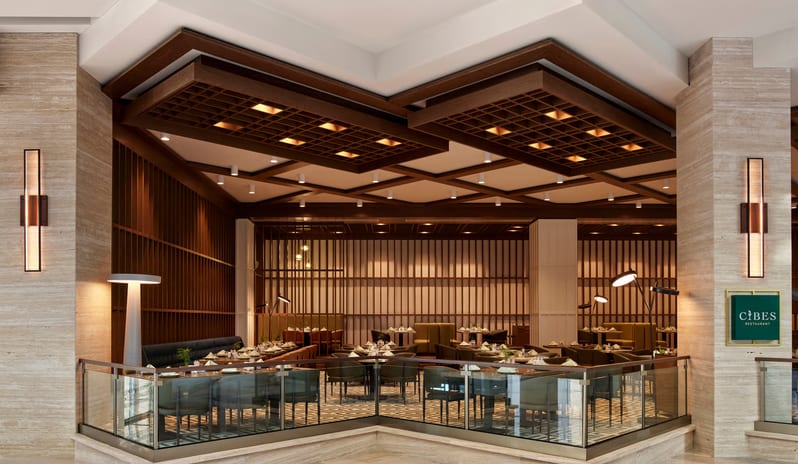 Cibes Restaurant International interior featuring an intricate dark wood lattice ceiling, travertine stone walls, and elegant dining tables set for service under warm ambient lighting at Anda Barut Collection by Kenwood Travel