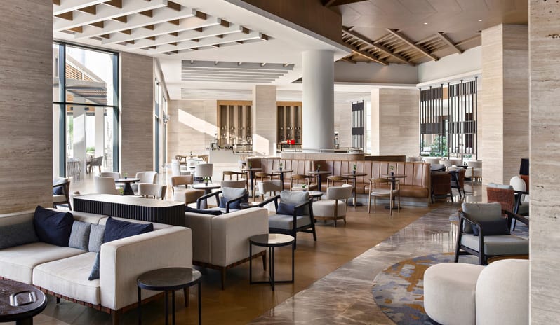 Olivae Patisserie lounge with neutral-toned armchairs, leather banquette seating, marble flooring, and contemporary architectural ceiling details at Anda Barut Collection by Kenwood Travel