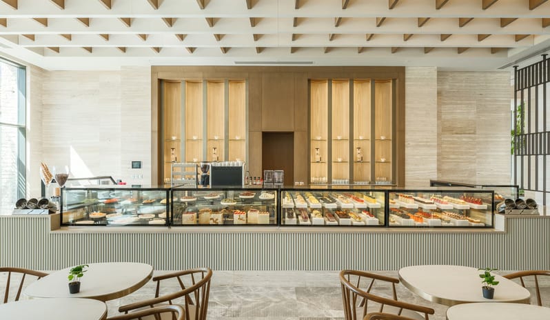 Olivae Patisserie featuring a long glass display counter filled with gourmet desserts, elegant wood-framed chairs, and a coffered ceiling with warm lighting at Anda Barut Collection by Kenwood Travel