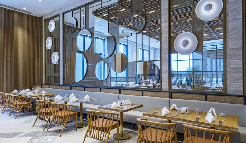 Zestful Bazaar Main Restaurantlight wood tables, spindle-back chairs, and a long gray banquette set against a decorative wall with circular mirrors and integrated disc lighting at Anda Barut Collection by Kenwood Travel