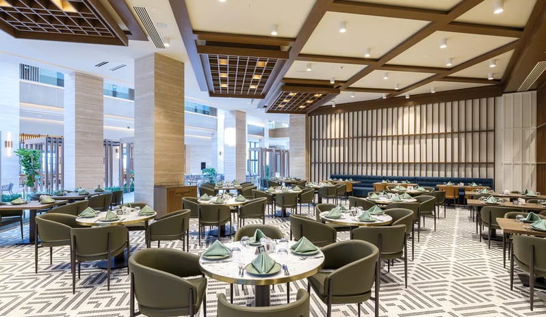 Cibes Restaurant International dining area with marble-topped tables and olive green chairs, featuring an architectural wood lattice ceiling and a view into a multi-story sunlit atrium at Anda Barut Collection by Kenwood Travel