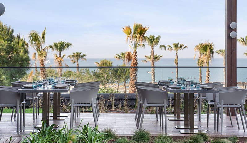 Cuisine24 restaurant terrace with grey chairs and stone tables overlooking a tropical beach with palm trees and the ocean at Voyage Belek Golf & Spa by Kenwood Travel