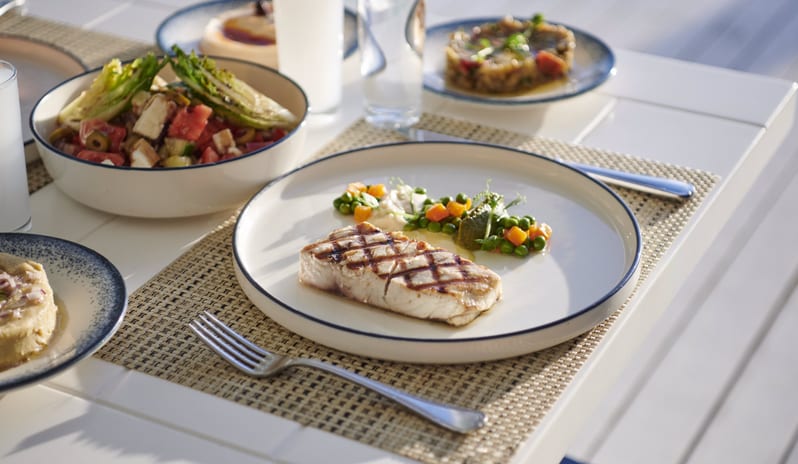 Blue Voyage Close-up of a grilled fish fillet with peas and carrots, served with a fresh Mediterranean salad and glasses of Raki on a white outdoor table at Voyage Belek Golf & Spa by Kenwood Travel