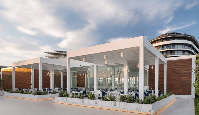 Blue Voyage Modern white glass restaurant pavilion with outdoor seating at a luxury beach resort with a circular hotel tower in the background at Voyage Belek Golf & Spa by Kenwood Travel