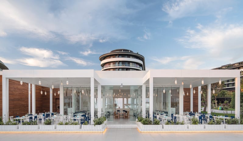 Blue Voyage Modern white glass restaurant pavilion with outdoor seating at a luxury beach resort with a circular hotel tower in the background at Voyage Belek Golf & Spa by Kenwood Travel