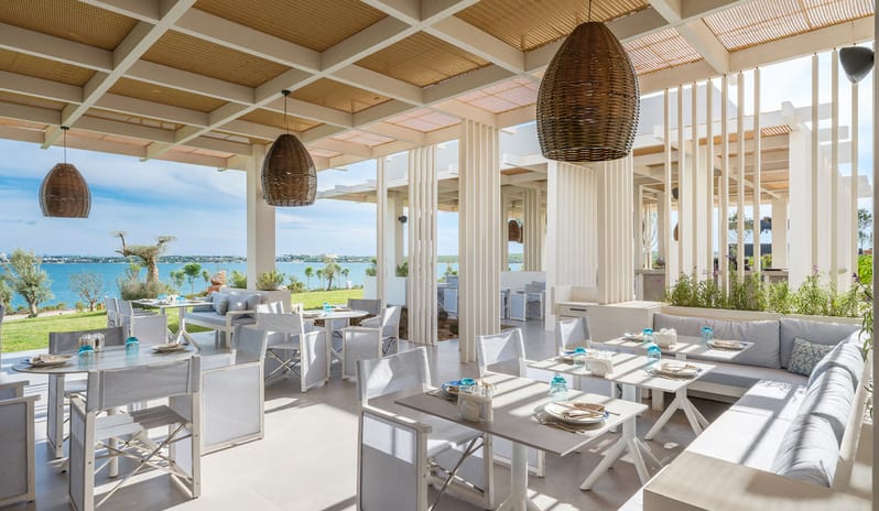 Fish Camp Restaurant outdoor restaurant terrace with white wooden dining tables, woven basket pendant lights, and a coffered ceiling offering a panoramic view of the blue ocean and coastline at Anda Barut Collection by Kenwood Travel