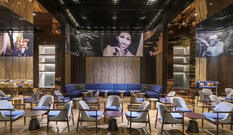 Soa Playhouse light blue armchairs, a long navy blue velvet sofa, and wooden herringbone flooring, decorated with large black-and-white jazz musician murals including Aretha Franklin at Anda Barut Collection by Kenwood Travel