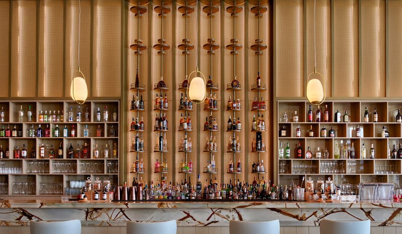 Soa Playhouse Ease Lobby Lounge Bar & Terrace sophisticated hotel bar featuring a white marble countertop with dark veining, light blue modern bar stools, and a warm gold-textured wall display of premium spirits at Anda Barut Collection by Kenwood Travel