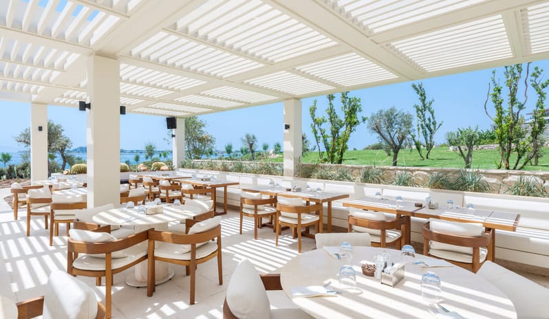 Cape Beach Lounge & Club South American patio with white louvered pergola, minimalist wooden furniture, and olive trees overlooking the blue Aegean Sea with a yacht in the distance at Cape Sounio at Anda Barut Collection by Kenwood Travel