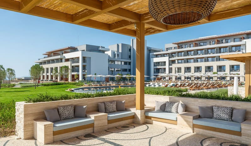 Mansion Restaurant International Kitchen lounge area with built-in stone seating, decorative mosaic pebble flooring, and a large wicker pendant light overlooking a luxury hotel complex and pool at Anda Barut Collection by Kenwood Travel