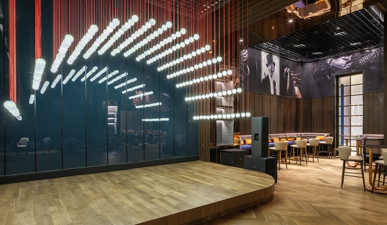 Soa Playhouse curved wooden performance stage at Sona Playhouse featuring a dramatic lighting installation of white glowing orbs suspended by red cables, set against dark wood paneling and jazz-themed wall art at Anda Barut Collection by Kenwood Travel