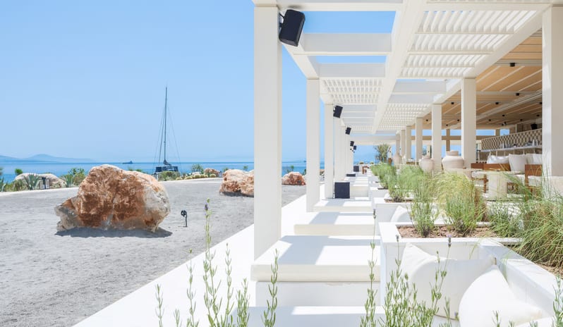 Cape Beach Lounge & Club South American white cushioned seating and minimalist pergolas under a clear blue sky, featuring coastal landscaping and a view of a sailboat on the sea at Cape Sounio at Anda Barut Collection by Kenwood Travel
