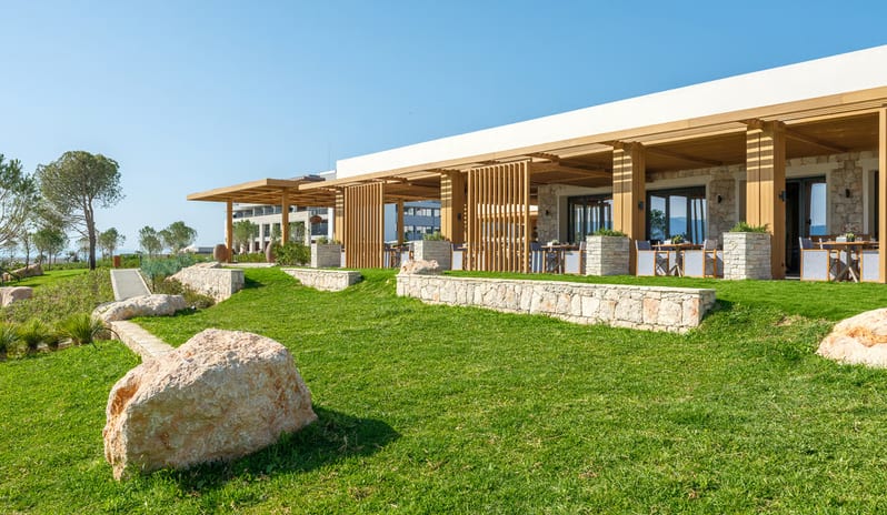 Mansion Restaurant International Kitchen featuring natural stone retaining walls, wooden pergolas, and a tiered green lawn with large decorative boulders under a bright sky at Anda Barut Collection by Kenwood Travel