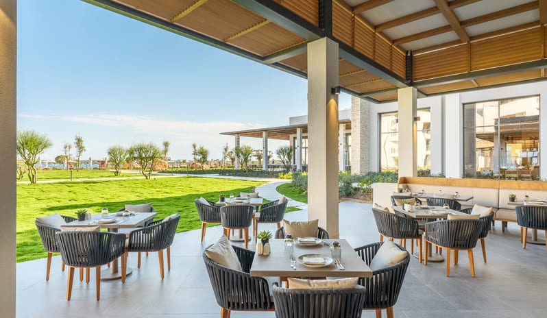 Kara Restaurant restaurant patio with elegant rope-back chairs, stone tables, and a wooden louvered ceiling overlooking a lush green lawn and garden at Anda Barut Collection by Kenwood Travel