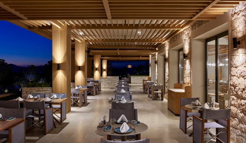 Mansion Restaurant International Kitchen outdoor restaurant terrace at night featuring warm pillar lighting, a modern wood-slat ceiling, and elegantly set tables with a twilight sky background at Anda Barut Collection by Kenwood Travel