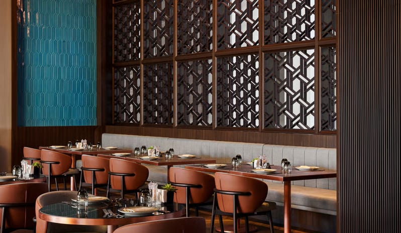 Kara Restaurant dining room featuring curved leather armchairs, dark wood tables, and a large decorative geometric wood lattice partition under warm pendant lighting at Anda Barut Collection by Kenwood Travel