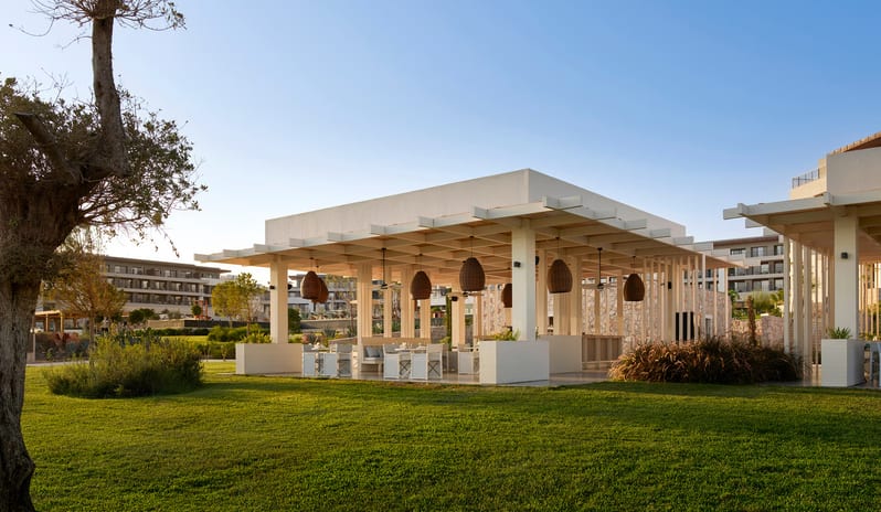 Fish Camp Restaurant open-air dining pavilion at a luxury resort featuring a wooden pergola ceiling, woven pendant lights, and white seating overlooking a lush green lawn at Anda Barut Collection by Kenwood Travel
