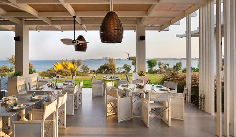 Fish Camp Restaurant outdoor restaurant terrace with white wooden dining tables, woven basket pendant lights, and a coffered ceiling offering a panoramic view of the blue ocean and coastline at Anda Barut Collection by Kenwood Travel