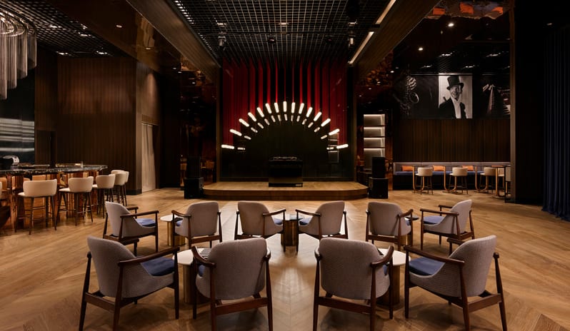 Soa Playhouse semicircular arrangement of gray armchairs on a herringbone wood floor, leading to a wooden performance stage with a sunburst lighting installation and a grand bar area to the left at Anda Barut Collection by Kenwood Travel