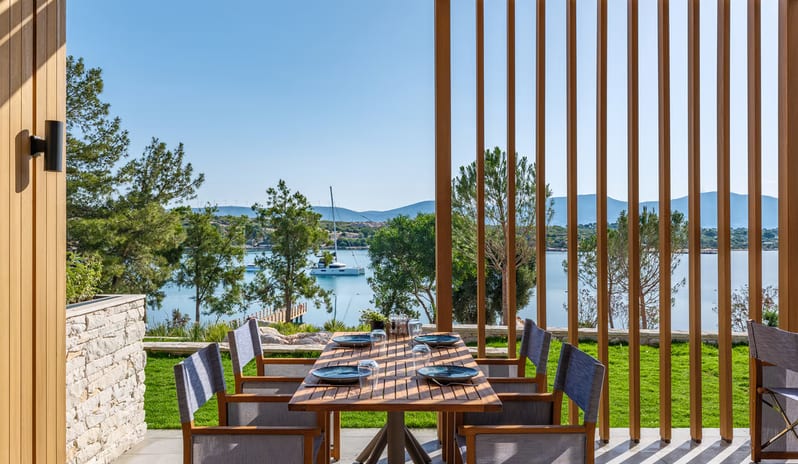 Mansion Restaurant International Kitchen outdoor dining table under a modern wooden slat pergola with a scenic view of a bay, featuring a white catamaran yacht and distant mountains at Anda Barut Collection by Kenwood Travel
