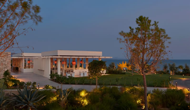 Fish Camp Restaurant evening view of a modern beachfront resort pavilion at dusk, featuring warm interior lighting, glowing garden path lights, and lush landscaping against a deep blue twilight sky at Anda Barut Collection by Kenwood Travel