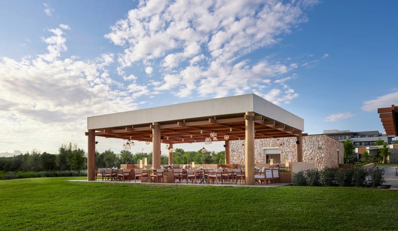 No Se Restaurant open-air restaurant pavilion with wooden pillars and a white flat roof, featuring elegant outdoor dining seating on a lush green lawn under a bright blue sky at Anda Barut Collection by Kenwood Travel