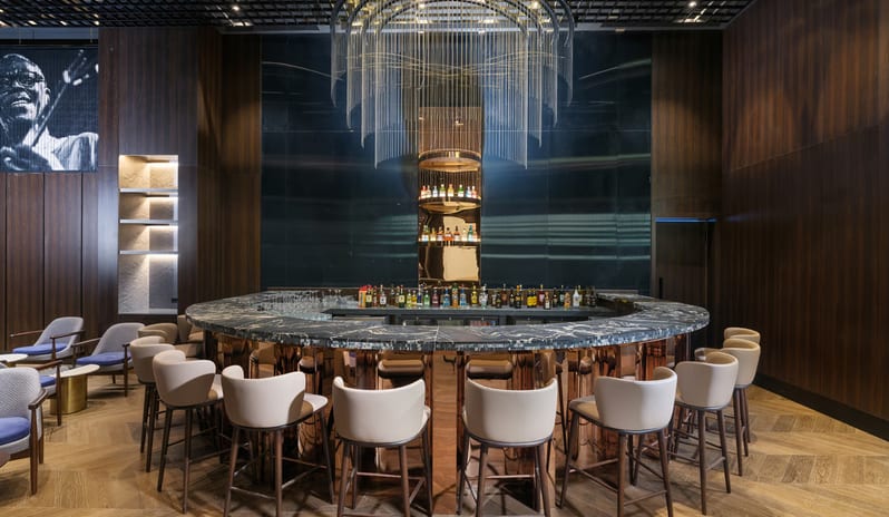 Soa Playhouse featuring a dark marble countertop, polished copper base, and cream-colored bar stools under a large modern chain chandelier at Anda Barut Collection by Kenwood Travel