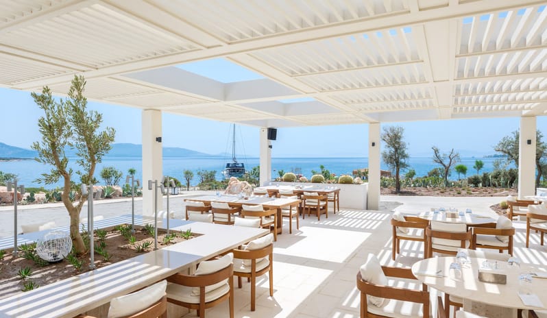 Cape Beach Lounge & Club South American patio with white louvered pergola, minimalist wooden furniture, and olive trees overlooking the blue Aegean Sea with a yacht in the distance at Cape Sounio at Anda Barut Collection by Kenwood Travel