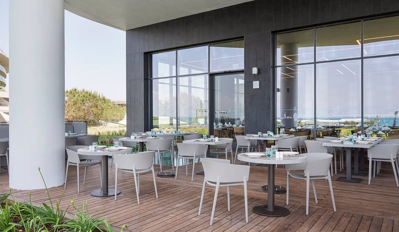 Degusto Adult Restaurant featuring gray modern chairs, round white tables, and floor-to-ceiling glass windows reflecting the resort landscape at Voyage Belek Golf & Spa by Kenwood Travel