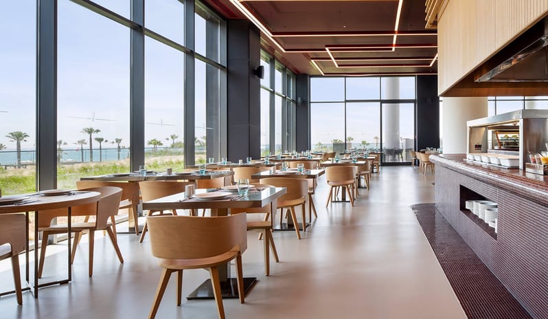 Degusto Adult Restaurant fmodern buffet area, sleek wooden furniture, and floor-to-ceiling windows with a scenic view of the Mediterranean coast at Voyage Belek Golf & Spa by Kenwood Travel