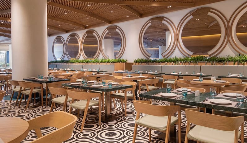 Degusto Main Restaurant green marble-topped tables, light wood chairs, and a striking black and white geometric patterned floor, with large circular wall mirrors and a slatted timber ceiling at Voyage Belek Golf & Spa by Kenwood Travel