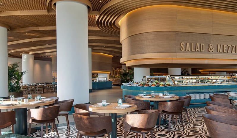 Degusto Main Restaurant curved wood-paneled buffet island, contemporary dark wood chairs, and an intricate geometric patterned floor under a slatted ceiling at Voyage Belek Golf & Spa by Kenwood Travel