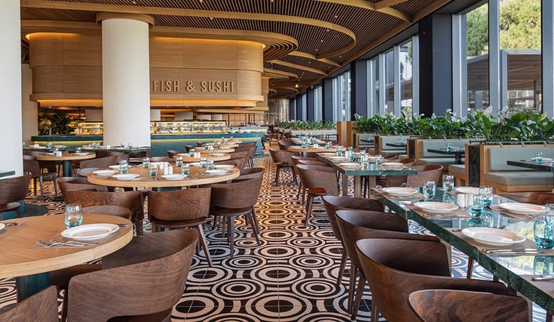 Degusto Main Restaurant green marble-topped tables, light wood chairs, and a striking black and white geometric patterned floor, with large circular wall mirrors and a slatted timber ceiling at Voyage Belek Golf & Spa by Kenwood Travel