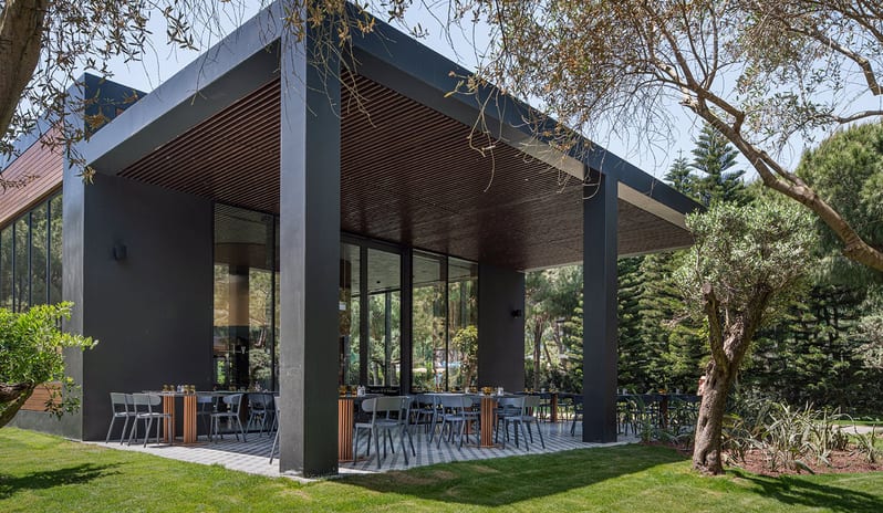 Voyage Belek Golf & Spa Blue Kebappa Turkish Restaurant modern black architecture, wooden slatted ceilings, and tables set on a tiled patio surrounded by green lawn and trees-Kenwood Travel at Voyage Belek Golf & Spa by Kenwood Travel