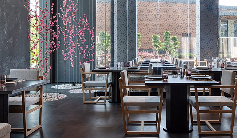 Wen Chinese Restaurant dark wood tables, grey upholstered chairs, and pink floral accents near large windowsat Voyage Belek Golf & Spa by Kenwood Travel