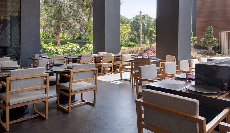 Wen Teppanyaki Japanese Restaurant outdoor dining area at Wen Teppanyaki Restaurant in Voyage Belek, featuring minimalist wooden chairs, dark tables, and a view of lush green gardens at Voyage Belek Golf & Spa by Kenwood Travel