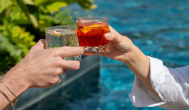Aqua Bar Two people clinking glasses of colorful cocktails by a blue swimming pool while playing a card game on a sunny day at Liberty Signa Hotel by Kenwood Travel