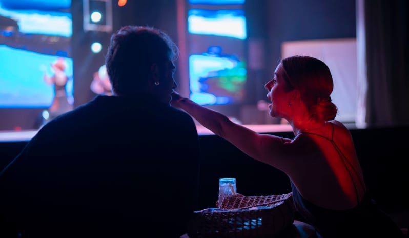 Arena Bar woman gesturing and talking to a man at a dimly lit esports arena bar with glowing blue gaming monitors in the background at Liberty Signa Hotel by Kenwood Travel