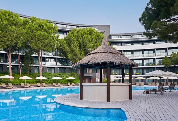 Garden Pool Bar luxury resort swimming pool with a central thatched-roof swim-up bar, surrounded by lounge chairs, white umbrellas, and tiered hotel balconies at Voyage Belek Golf & Spa by Kenwood Travel