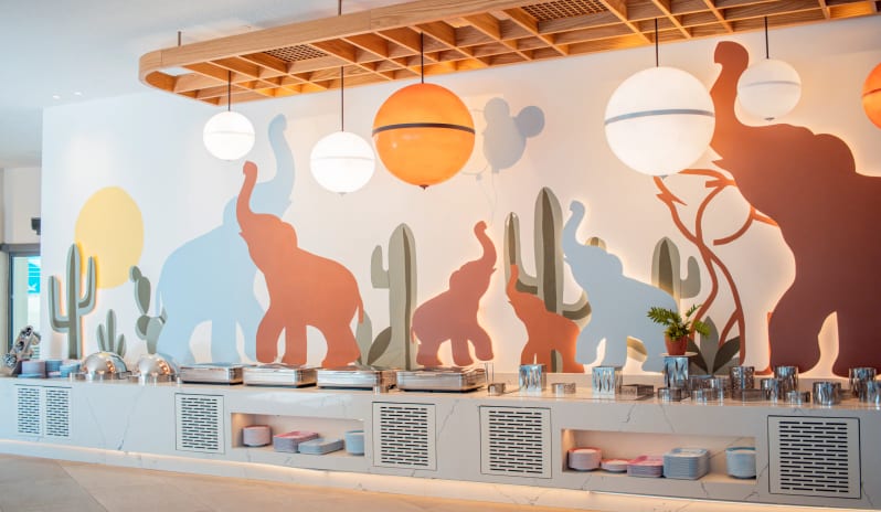 Myra Main Restaurant breakfast buffet station at Primo Piatto in Disney's Riviera Resort featuring a colorful mural of elephants, cacti, and spherical pendant lighting at Liberty Fabay by Kenwood Travel