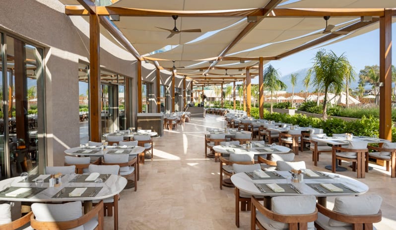 Myra Main Restaurant & Kids Buffet outdoor restaurant terrace with marble-top tables, wooden chairs, and a white fabric canopy overlooking palm trees and mountains at Liberty Signa Hotel by Kenwood Travel