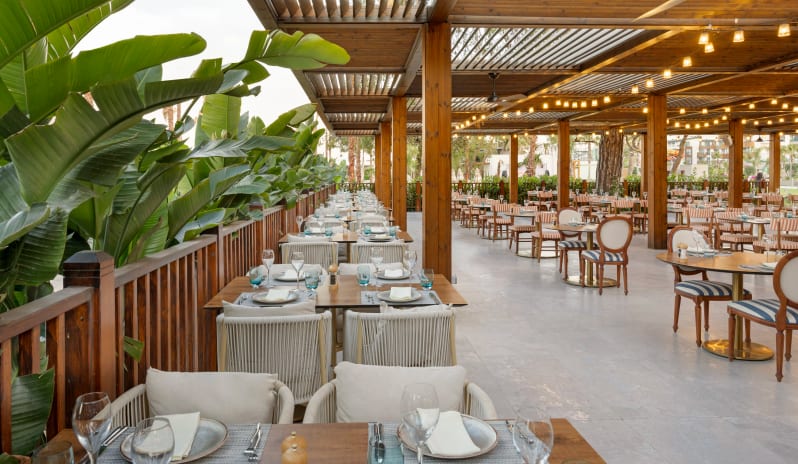 Okeanos A’la Carte Restaurant featuring wooden pergola roofing, hanging bistro lights, elegant table settings, and lush tropical greenery at Liberty Signa Hotel by Kenwood Travel
