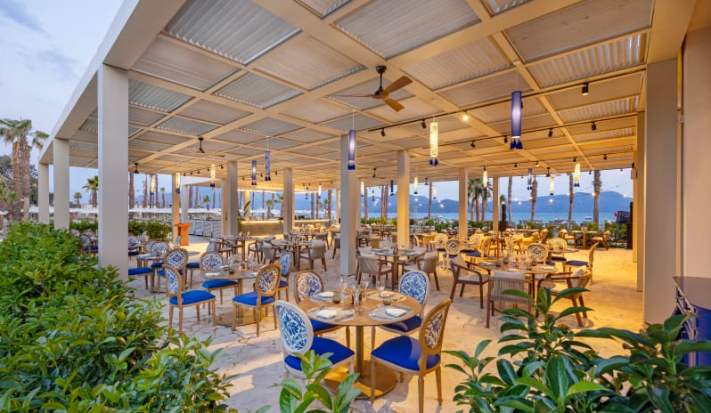 Tatre A’la Carte Restaurant open-air Mediterranean restaurant with blue and white patterned chairs, wooden tables, and hanging lanterns under a louvered pergola at dusk at Liberty Signa Hotel by Kenwood Travel